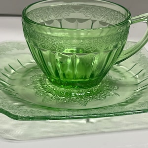 Può includere: Un set composto da una tazza da tè e un piattino quadrato abbinato in vetro verde chiaro traslucido. La tazza presenta una fascia orizzontale con motivi floreali in rilievo vicino al bordo e scanalature verticali alla base. Il piattino quadrato mostra un motivo floreale centrale.