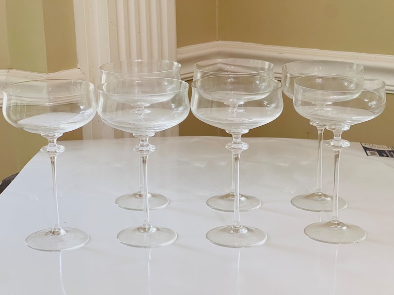 May include: Six clear glass champagne coupes on a white surface. The coupes are all the same size and shape, and they are arranged in a row.