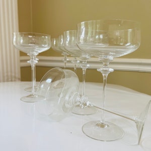 May include: A set of six clear glass champagne flutes with a wide bowl and a long stem. The flutes are arranged in a row on a white surface.