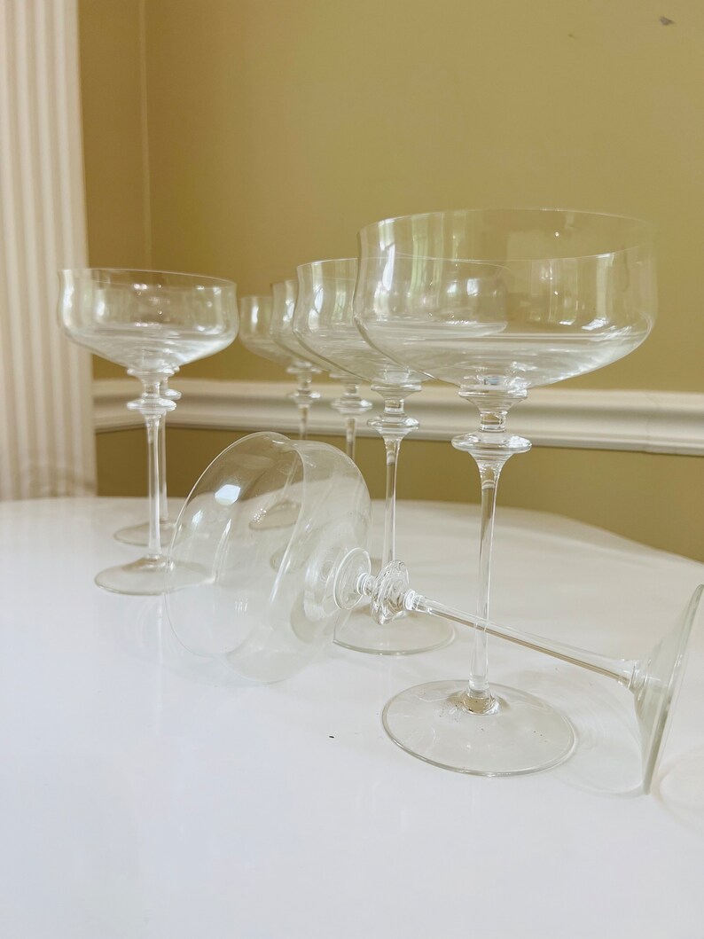 May include: A set of six clear glass champagne flutes with a wide bowl and a long stem. The flutes are arranged in a row on a white surface.