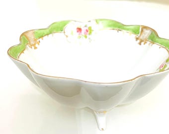 May include: A white porcelain bowl with a green and gold rim and floral design. The bowl has a scalloped edge and three small feet.