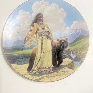 Puede incluir: Un plato de cerámica decorativo que presenta a una mujer nativa americana con ropa tradicional de pie con un oso marrón en un paisaje montañoso. El plato está decorado con un cielo azul y blanco, hierba verde y una cascada. La firma del artista, "David Wright", es visible en la esquina inferior derecha.
