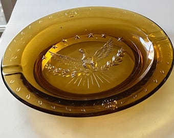 May include: Amber-colored glass ashtray with an oval shape. The center features an embossed design of an eagle with outstretched wings, surrounded by stars and a laurel wreath. The rim has a decorative pattern.