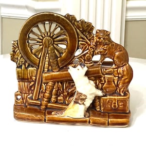 Puede incluir: Un macetero de cerámica marrón y blanco con forma de rueda de hilar con una figura de gato y perro. El macetero tiene un aspecto rústico y vintage.