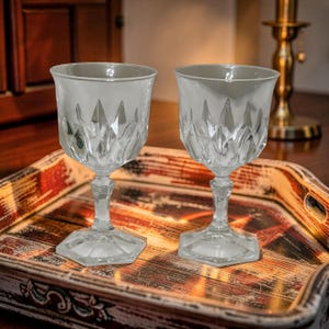 May include: Two clear, cut-glass goblets with a diamond pattern, resting on a decorative tray. The goblets have a hexagonal base and a detailed stem. The tray has a distressed, rustic design with red and brown tones.