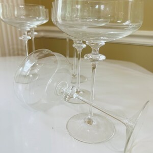 May include: Four clear glass champagne coupes with a delicate stem and a wide bowl. The glasses are arranged on a white surface.