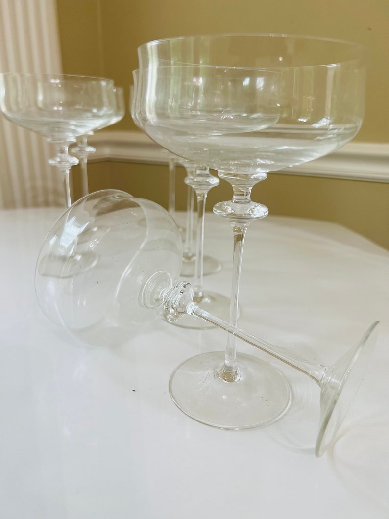 May include: Four clear glass champagne coupes with a delicate stem and a wide bowl. The glasses are arranged on a white surface.