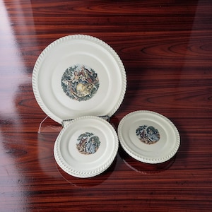 May include: A set of three antique white ceramic plates with a decorative rope-like edge. Each plate features a central image of a historical scene with figures in colorful attire. The plates are arranged on a dark wood surface.