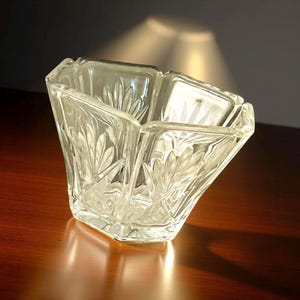 May include: Clear, square-shaped glass bowl with a leaf pattern. The bowl has a unique, angled design and sits on a wooden surface. The glass reflects light, creating highlights and shadows. A decorative piece for the home.