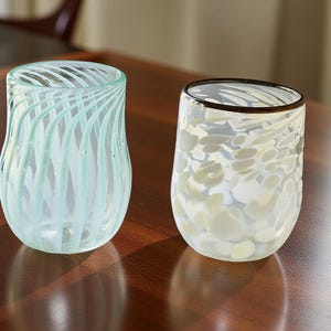 May include: Two decorative glass tumblers. One features a light blue swirl pattern, the other a white and yellow speckled design with a dark rim. Both are displayed on a dark wooden surface.