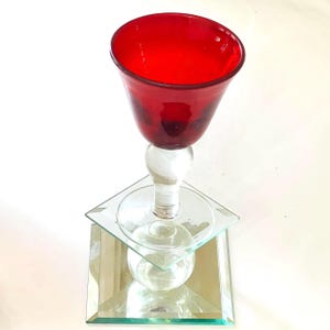 May include: A decorative glass object featuring a vibrant red bowl-shaped top supported by a clear glass stem. The stem rests on a square, clear glass base, which is positioned atop a square mirrored platform, creating a layered, reflective effect.