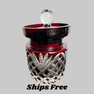 Op de afbeelding: Een rode glazen pot met een helder glazen deksel en ingewikkelde geslepen glasontwerpen. De pot heeft een donkerrode basis en een helder glazen bovenkant met een decoratief ontwerp.