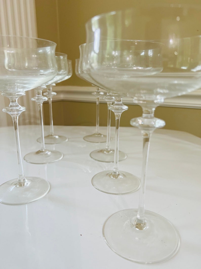 May include: A set of eight clear glass champagne flutes with a wide bowl and a long, thin stem. The flutes are arranged in a row on a white surface.