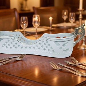 Puede incluir: Una fuente de porcelana blanca con un diseño calado y asas doradas. La fuente está sobre una mesa de madera con cubiertos y velas, lo que sugiere un ambiente de comedor formal.