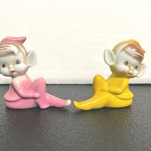 May include: Two ceramic elf figurines, one in pink and one in yellow, with painted details and brown hair. The elves are sitting with their legs crossed and their hands clasped in front of them.