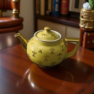 Puede incluir: Una tetera amarillo pálido con detalles dorados y tapa. La tetera está decorada con patrones dorados de flor de lis. El asa, el pico y el pomo son dorados. La tetera está sobre una superficie de madera oscura.