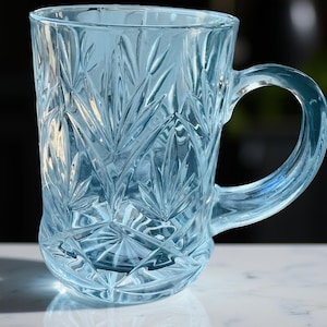 May include: A clear, light blue glass mug with a handle. The mug features a detailed, textured pattern of diamond shapes and leaf-like designs. The glass is transparent, allowing light to pass through, and the mug sits on a white surface.