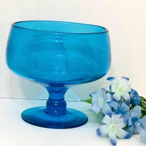 May include: A vibrant turquoise glass pedestal bowl with a rounded top and a sturdy base. The bowl is a bright, solid color and is displayed with a sprig of blue and white flowers. This decorative bowl is ideal for serving.