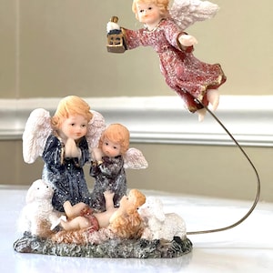 Pode incluir: Um conjunto de figuras de presépio natalino com três anjos e dois cordeiros. O anjo do centro está ajoelhado e rezando, enquanto o anjo da esquerda segura uma lanterna. O anjo da direita está voando com uma auréola branca e segurando uma lanterna. O menino Jesus está deitado em uma manjedoura com dois cordeiros ao seu lado.
