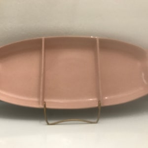 May include: A pale pink ceramic serving dish with three divided sections. The dish has a curved shape and a gold metal stand.