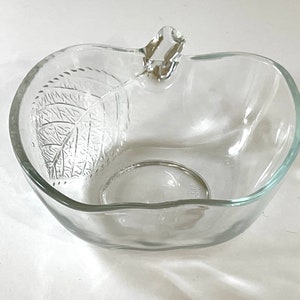 May include: A clear glass bowl shaped like an apple with a leaf design. The bowl has a handle on the top.