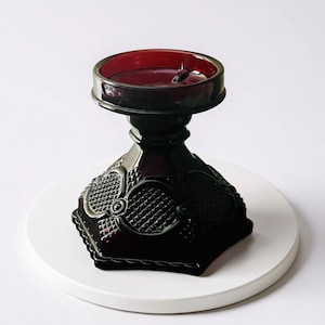 May include: A dark red glass candle holder with a decorative, geometric pattern. The candle holder has a wide, circular top and a pedestal base. The base is square with rounded edges. The candle holder is sitting on a white surface.