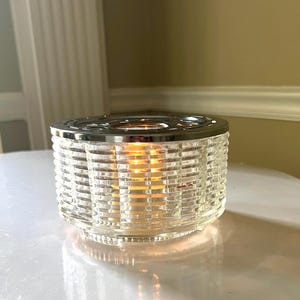 May include: A clear glass candle holder with a silver metal top. The holder has a grid pattern and is lit by a candle inside.