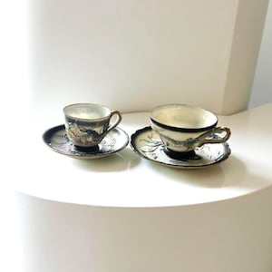 May include: Two matching teacups and saucers. The cups and saucers are black with a detailed design of a dragon. The cups have gold handles and a white interior. The saucers have a scalloped edge.