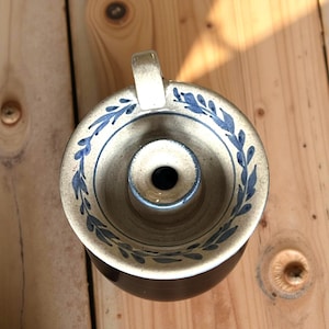 May include: A ceramic candle holder with a cream exterior and a dark brown base. The rim is decorated with blue leaf patterns. It has a small handle and a central opening for a candle.