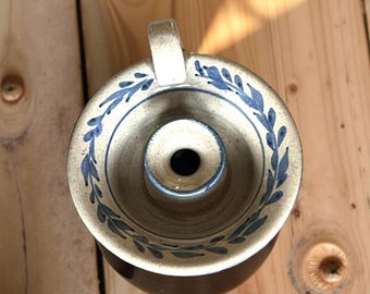 Könnte beinhalten: Ein handgefertigter Kerzenhalter aus Keramik mit cremefarbener Außenseite und dunkelbraunem Sockel. Der Rand ist mit blauen Blättermustern verziert. Er hat einen kleinen Griff und eine zentrale Öffnung für eine Kerze.