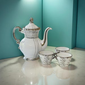 May include: A white porcelain tea set with silver detailing. The set comprises a teapot with a decorative lid and three teacups. The teapot features a curved spout and handle. The teacups and teapot are adorned with a floral design.