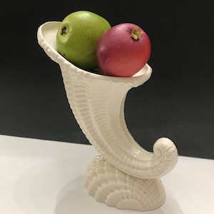 May include: A white ceramic cornucopia vase with a textured surface, holding a green apple and a red apple.