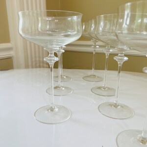 May include: Set of six clear glass champagne flutes with a wide bowl and a long, thin stem. The flutes are arranged in a semi-circle on a white surface.