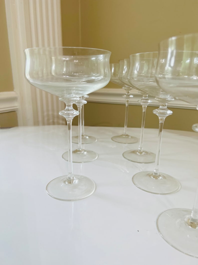 May include: Set of six clear glass champagne flutes with a wide bowl and a long, thin stem. The flutes are arranged in a semi-circle on a white surface.