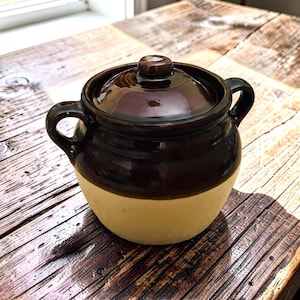 May include: A two-handled ceramic crock with a lid, featuring a glossy dark brown glaze on the upper portion and a matte cream-colored base. The pot has a rounded body, a short neck, and a small knob handle centered on the circular lid.