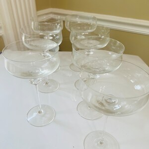 May include: Set of eight clear glass champagne coupes with a wide bowl and a long stem. The coupes are arranged in a row on a white surface.