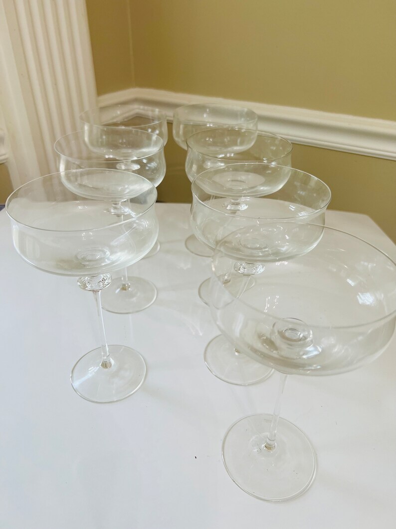 May include: Set of eight clear glass champagne coupes with a wide bowl and a long stem. The coupes are arranged in a row on a white surface.