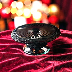 May include: A black glass cake stand featuring a circular top with a radiating sunburst pattern and a decorative embossed border. The pedestal base is also made of black glass with a textured, patterned design and a smooth, circular foot.