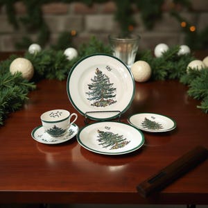 May include: A white ceramic Christmas dinnerware set with a green border. The set includes a teacup and saucer, a small plate, a medium plate, and a large plate. Each piece features a detailed illustration of a decorated Christmas tree.