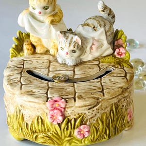 Puede incluir: Una caja de música de cerámica con dos gatitos jugando con un paño blanco. La caja está decorada con hierba verde, flores rosas y un camino de ladrillos marrones. Los gatitos están pintados en tonos de naranja, blanco y gris.