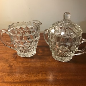 Può includere: Un set di lattiera e zuccheriera in vetro trasparente. Entrambi i pezzi presentano un motivo a diamante. La lattiera ha un manico e un bordo smerlato. Lo zuccheriera ha un coperchio con un piccolo pomello e due manici. Il set è su una superficie di legno.