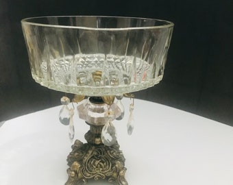 May include: A clear glass bowl with a decorative gold metal base and crystal-like drops hanging from the base. The bowl has a fluted design and is perfect for serving snacks or holding decorative items.
