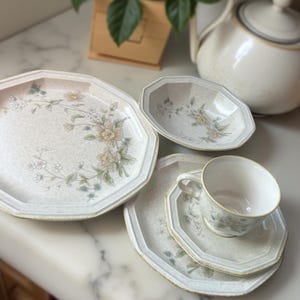 May include: A vintage floral-patterned tea set featuring a large octagonal plate, a bowl, a teacup with saucer, and a teapot. The set has a cream background with yellow, blue, and green floral designs. The teapot is white with a gold trim.