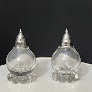 Puede incluir: Un juego de dos saleros y pimenteros de vidrio transparente con tapas plateadas y una base texturizada con forma de burbujas.