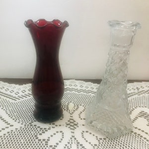 May include: Two glass vases, one is red and the other is clear with a textured design. Both vases are on a white lace tablecloth.