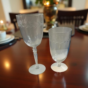 May include: Two clear glass goblets with etched leaf patterns. One has a tall stem and a rounded bowl, the other is a shorter tumbler. Both are on a dark wooden table, suggesting a dining setting.