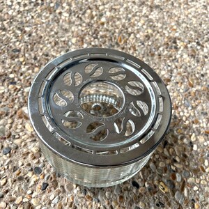 May include: A silver metal tea infuser with a glass base and a decorative design of leaves. The infuser has a handle on the side.