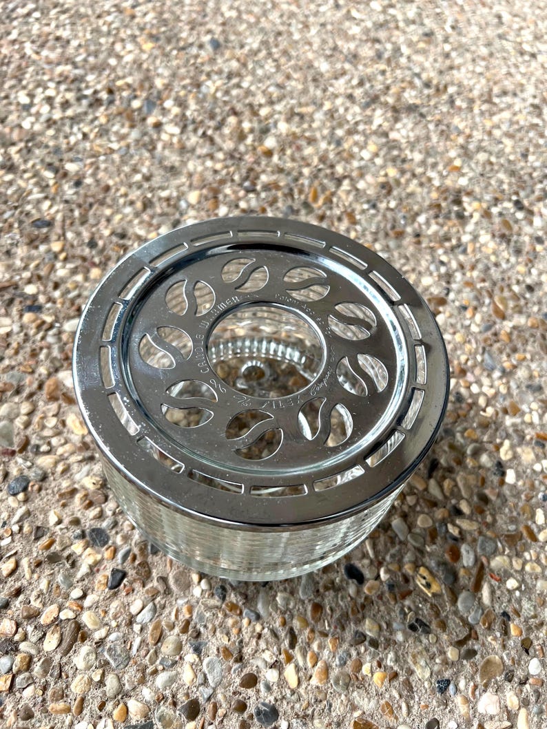 May include: A silver metal tea infuser with a glass base and a decorative design of leaves. The infuser has a handle on the side.