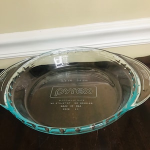 Op de afbeelding: Een heldere Pyrex taartvorm met een turquoise rand en handvatten. De schaal heeft een diameter van 24 cm en het Pyrex-logo, met tekst die aangeeft dat het magnetronbestendig is en niet geschikt voor kookplaat of grill. Gemaakt in de VS.