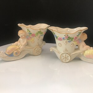 May include: A pair of vintage porcelain cherub planter vases. Each vase features a cherub riding a snail-shaped chariot with gold accents and floral details. The vases are in a cream color with pink, blue, and green accents.